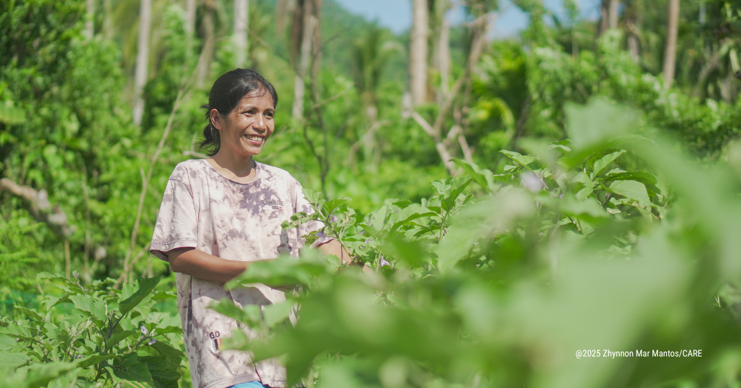 A farmer stands in a green vegetable farm surrounded by leafy crops, with trees and hills visible in the background.