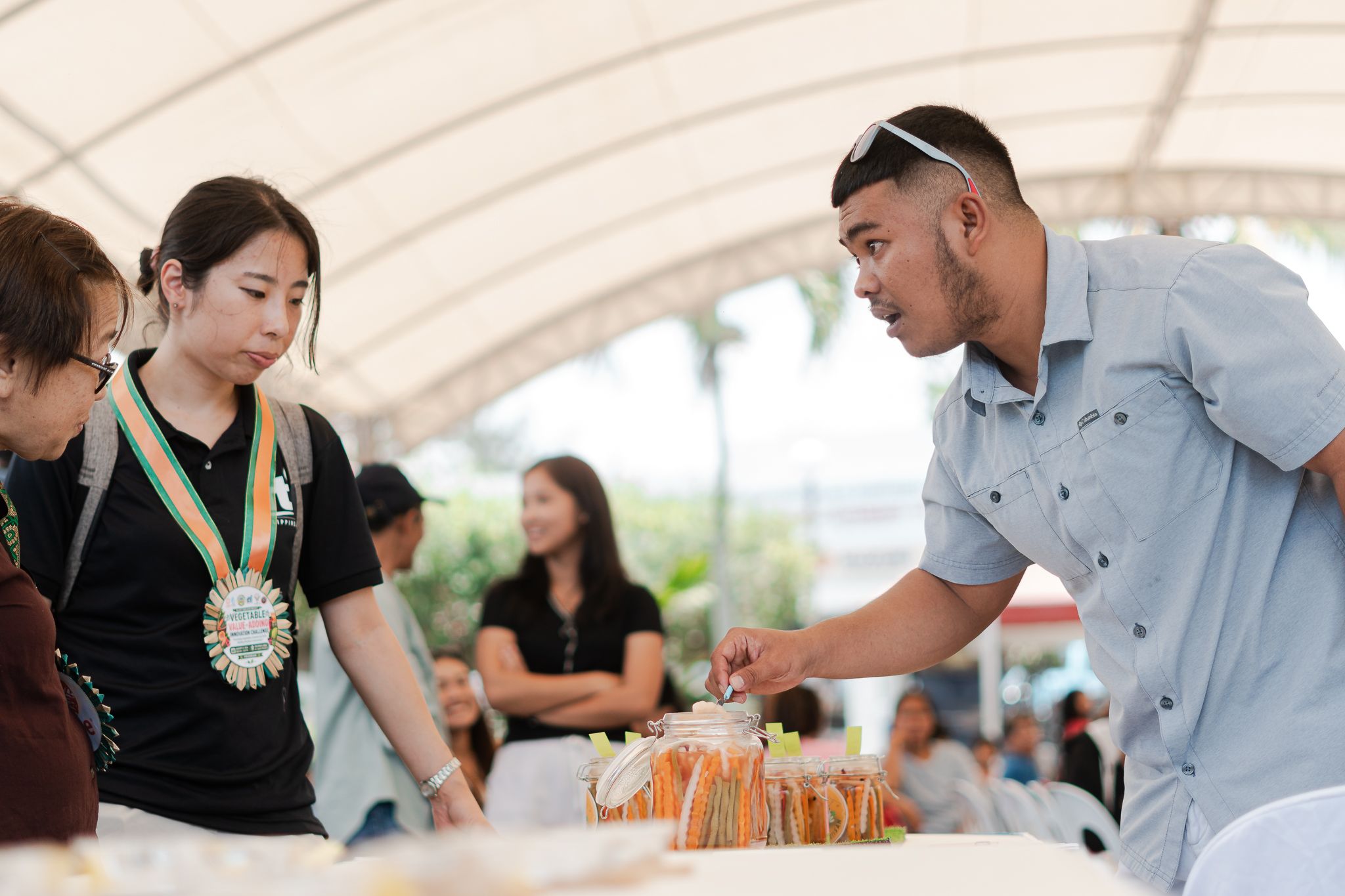 Judges stand at a table covered with homemade vegetable products as a participant offers them a taste.