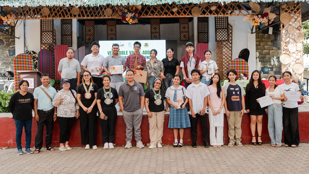 A large group of participants and guests pose together outdoors, several holding certificates. 